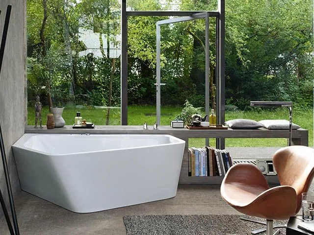 Freistehende weiße Badewanne vor einer großen Fensterfront mit Blick ins Grüne.