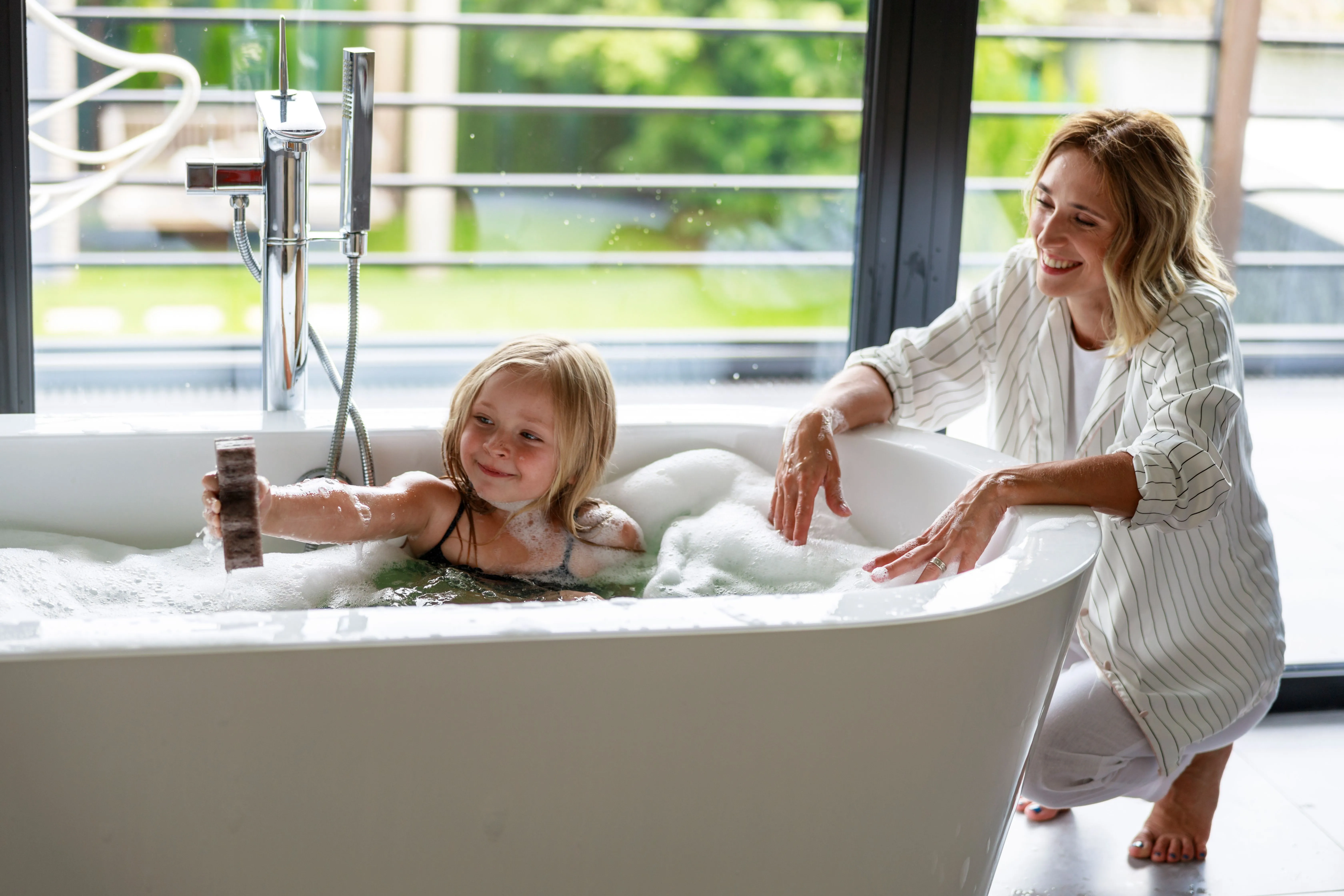 Mutter und Tochter an einer freistehenden Badewanne mit bodenstehender Armatur vor einer großen Fensterfront.
