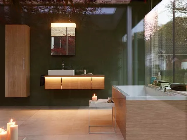 Stimmungsvoll beleuchtetes Bad mit Holz-Waschtischmöbel, Hochschrank und Einbauwanne an einer Glasfront zum Garten.