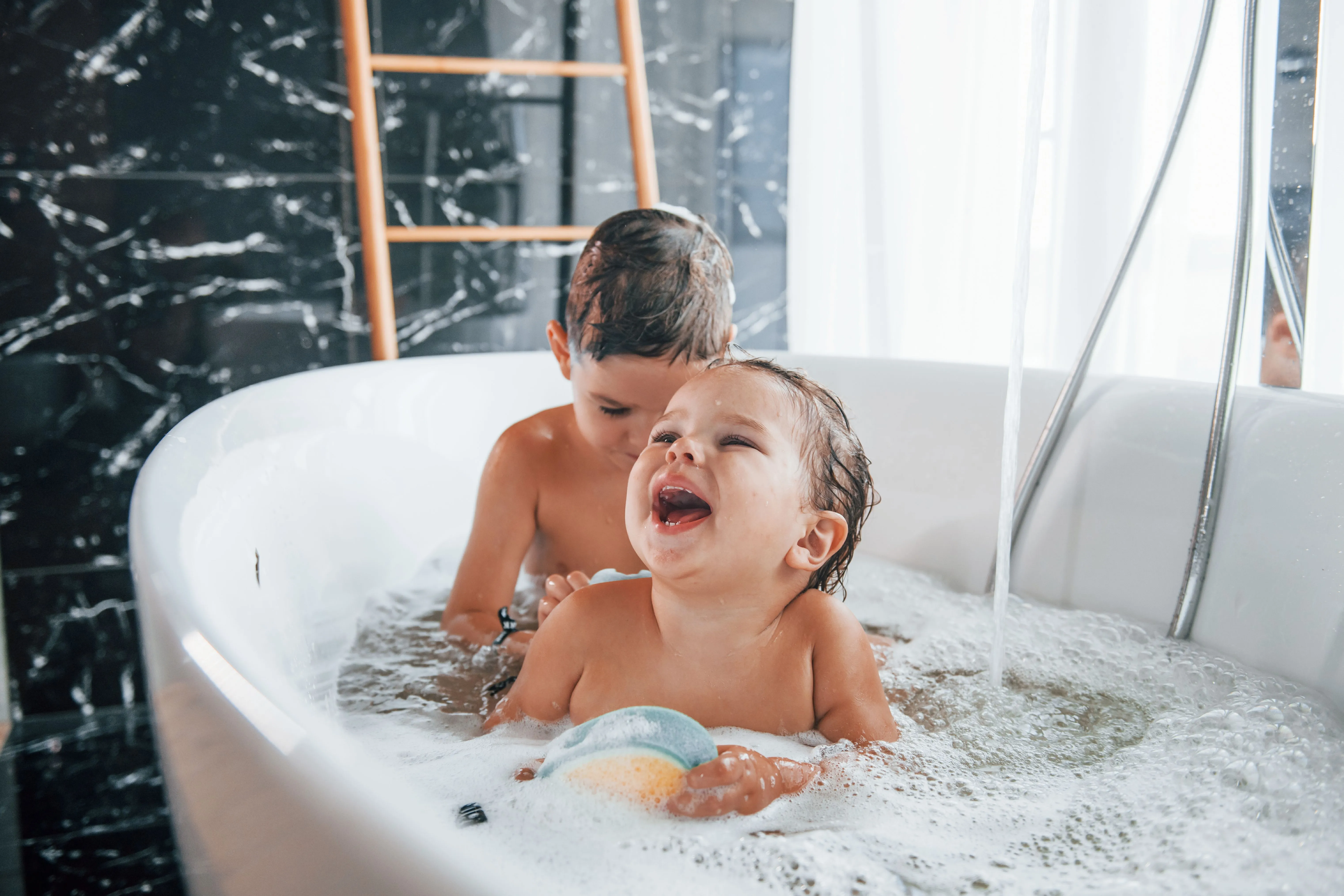 Zwei Kinder baden vergnügt mit Schaum in einer freistehenden Badewanne vor einer dunklen Marmorwand.