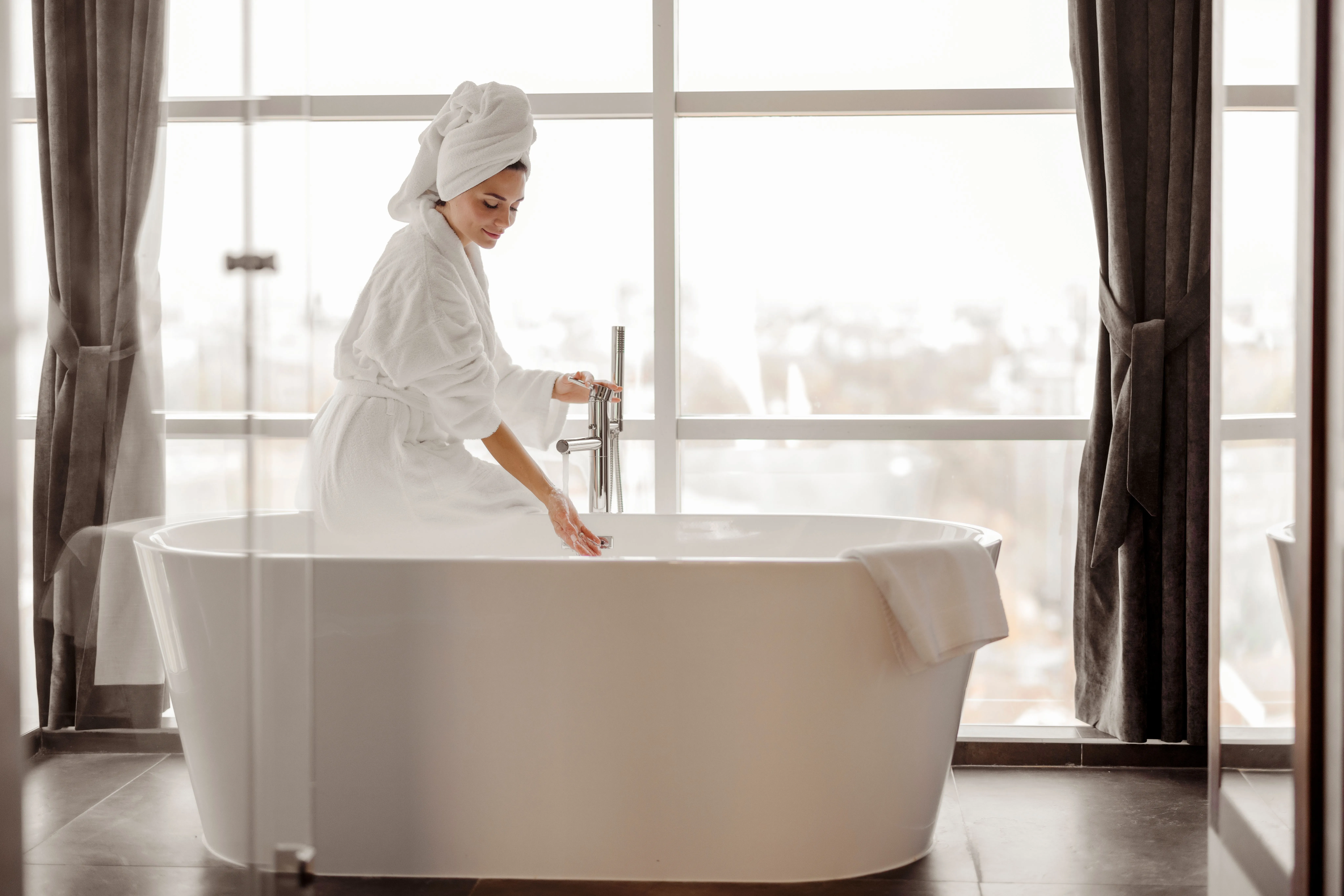 Frau im Bademantel mit Handtuchturban lässt Wasser in eine freistehende Badewanne vor großer Fensterfront ein.