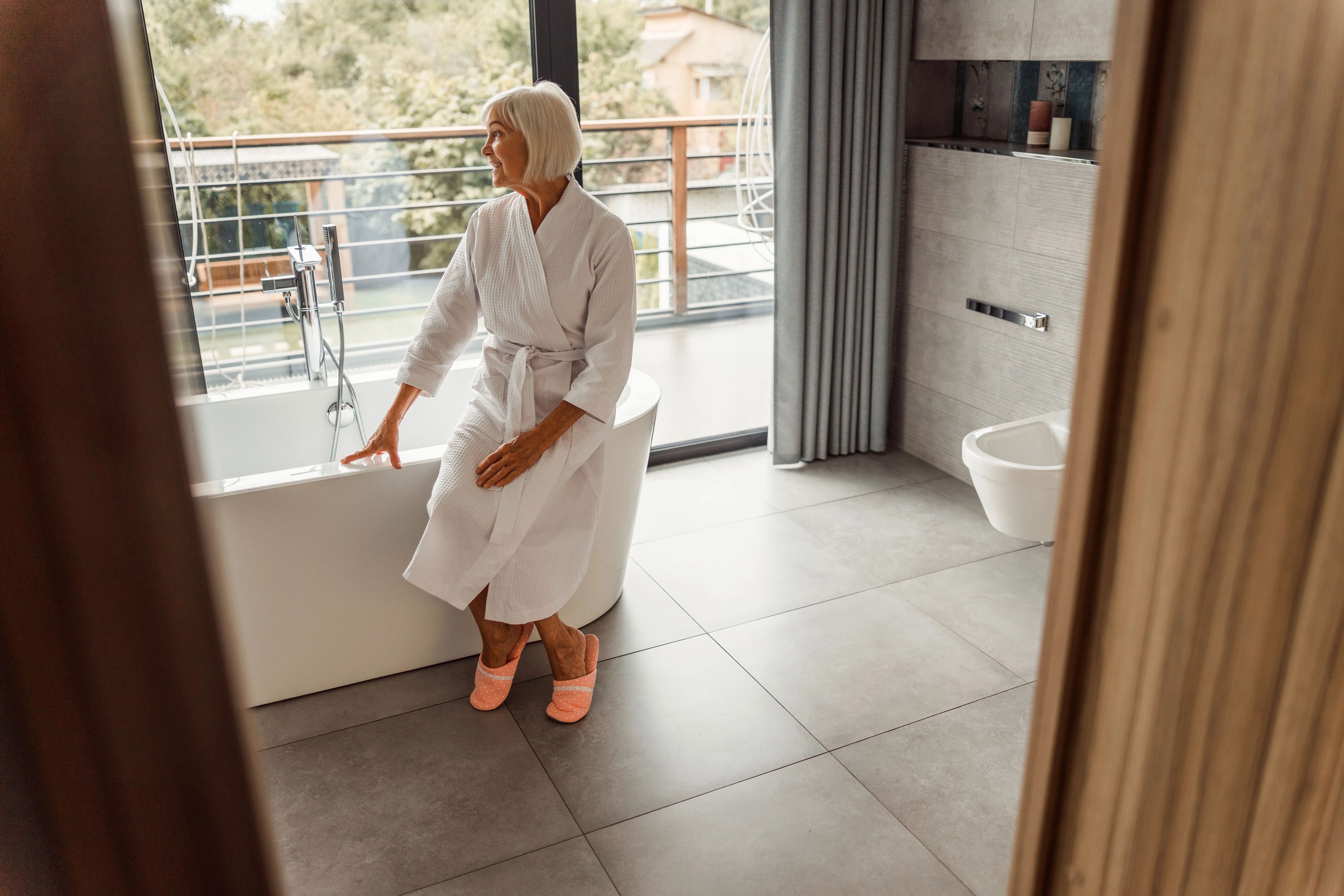 Seniorin im Bademantel sitzt entspannt auf dem Rand einer freistehenden Badewanne in einem modernen Bad mit Fensterfront.
