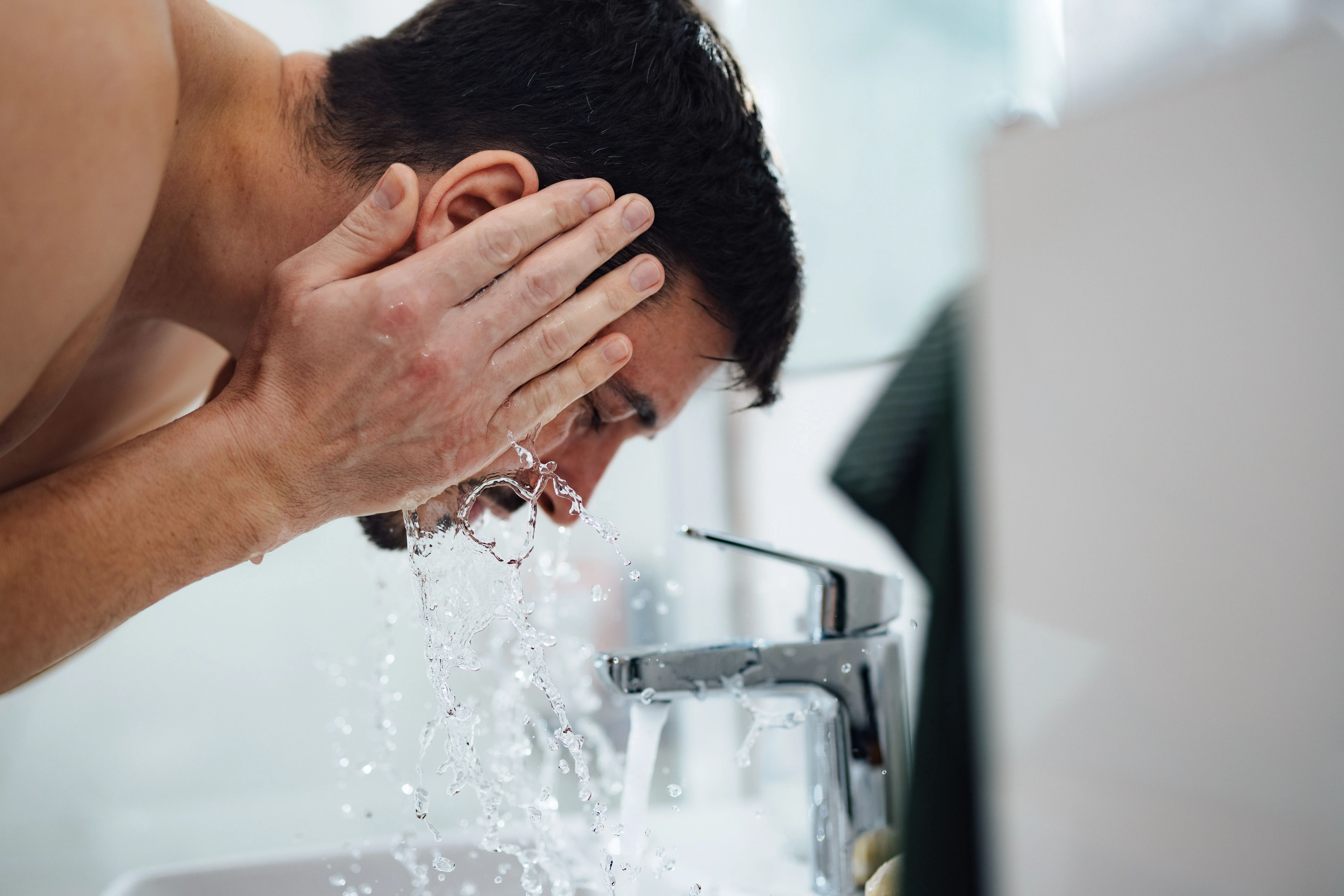 Mann wäscht sich am Waschbecken mit fließendem Wasser aus einer verchromten Armatur das Gesicht.