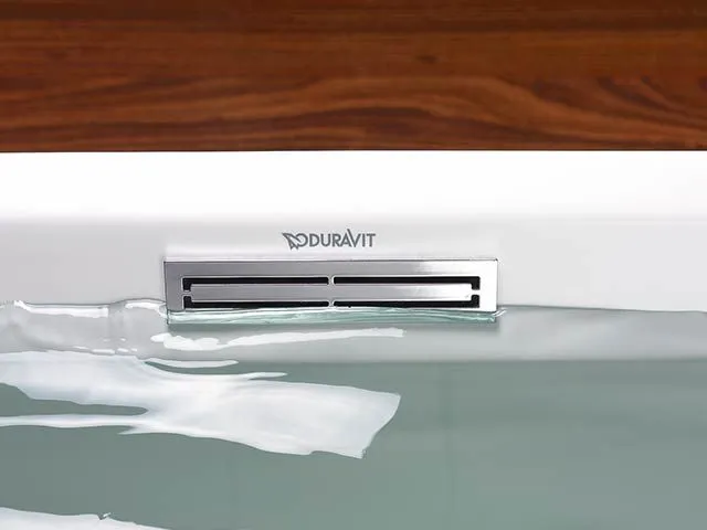 Detail einer gefüllten Badewanne mit rechteckigem Überlauf vor einer Holzverkleidung.
