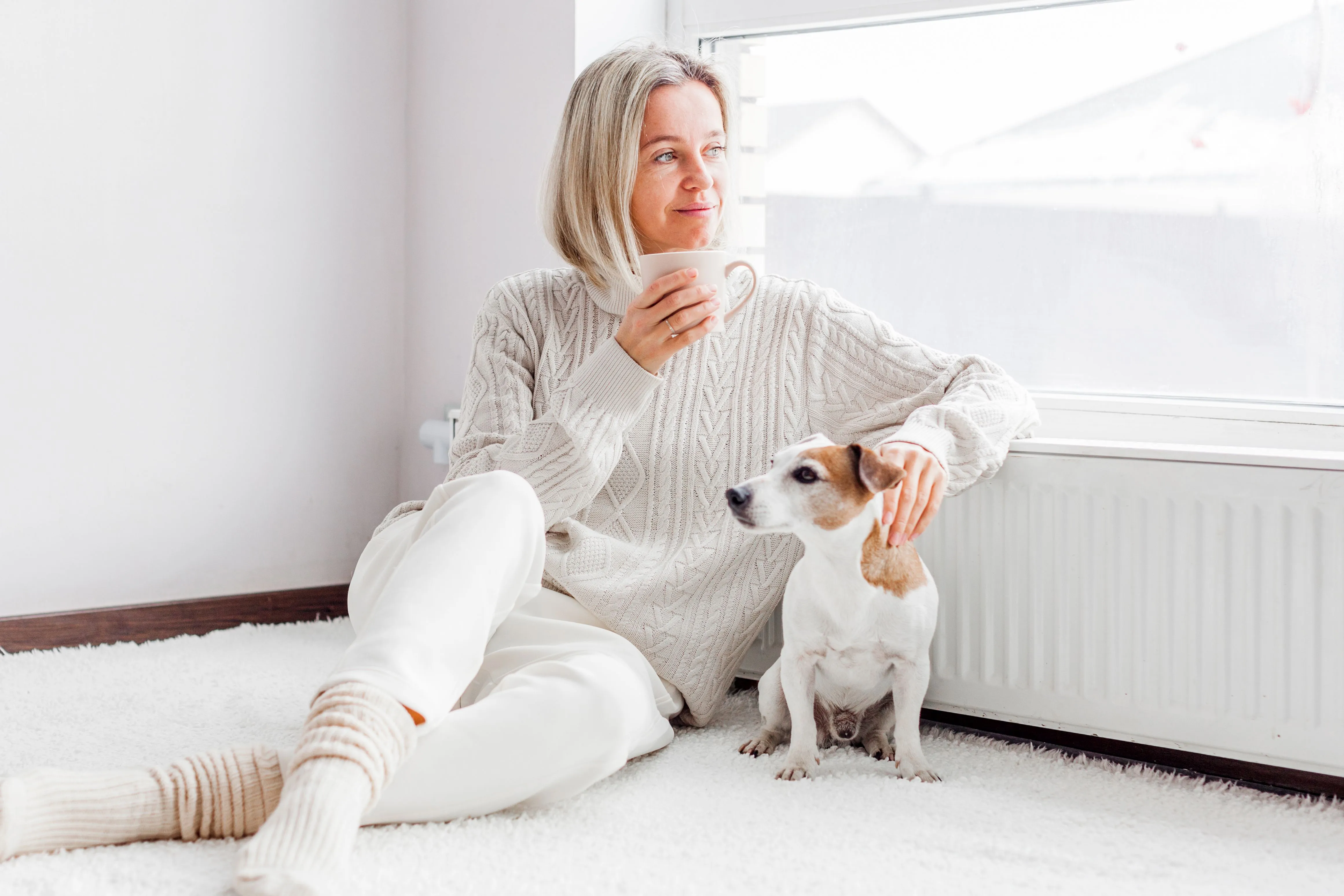 Frau mit Teetasse sitzt mit ihrem Hund entspannt auf dem Boden an einem warmen Heizkörper unter dem Fenster.