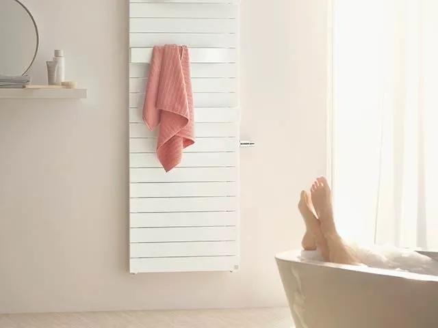 Weißer vertikaler Designheizkörper mit Handtuch neben einer sonnigen Badewanne.