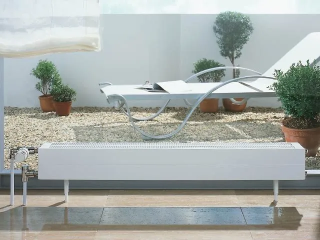Flacher weißer Konvektor-Heizkörper auf einer sonnigen Terrasse mit Liegestuhl und Kübelpflanzen.