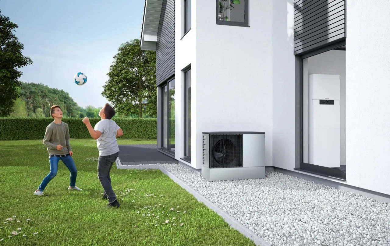 Luft-Wasser-Wärmepumpe am Einfamilienhaus, während zwei Kinder im Garten Fußball spielen.