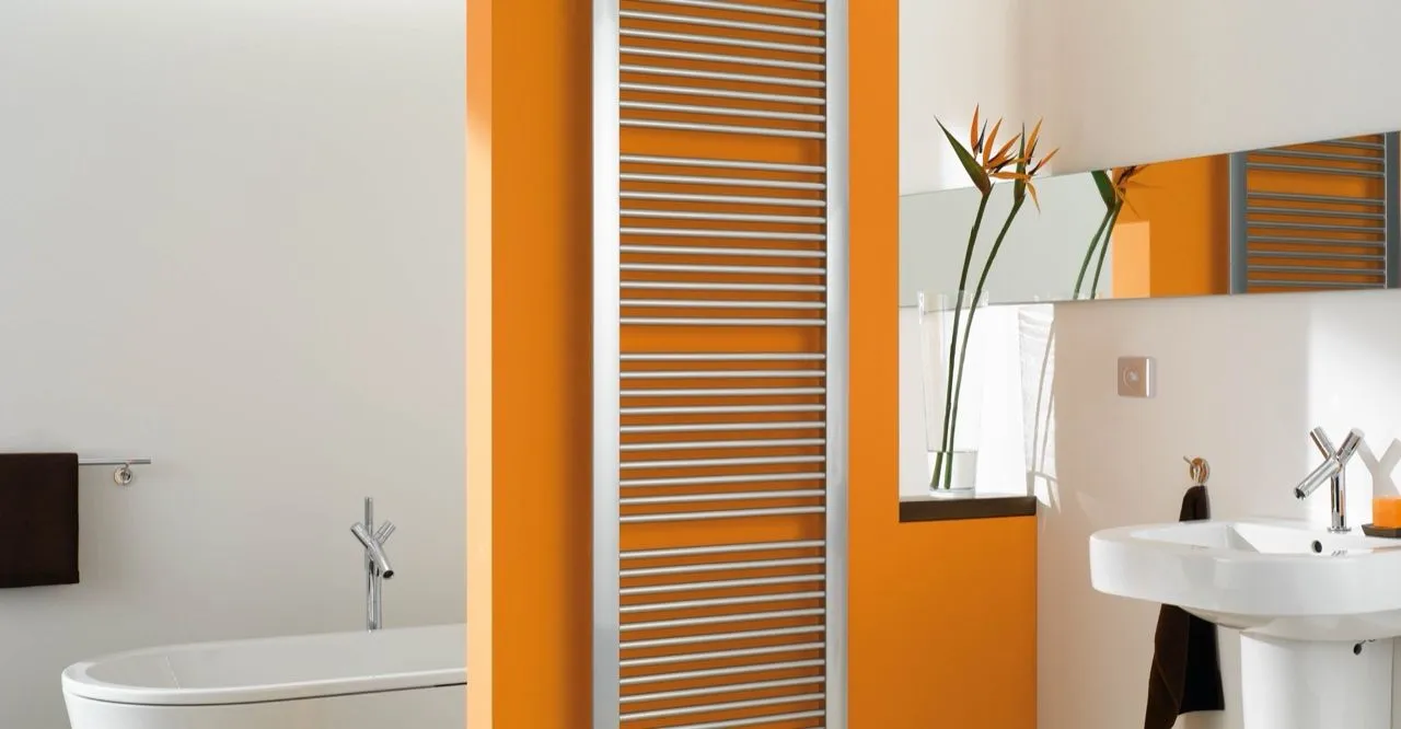 Verchromter Handtuchheizkörper an einer orangefarbenen Wand in einem hellen Bad mit Badewanne und Waschtisch.