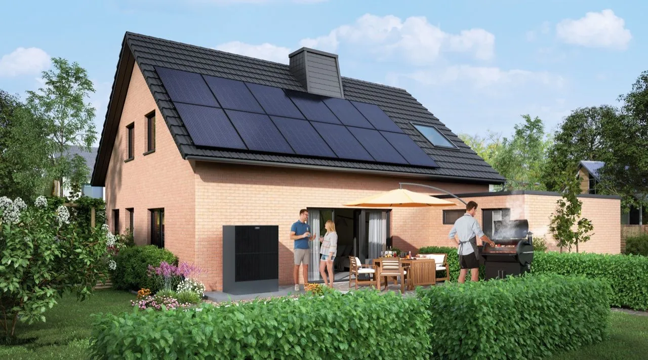 Modernes Backsteinhaus mit Photovoltaikdach; im Garten genießt eine Familie die Terrasse, an der Hauswand steht der Stromspeicher.
