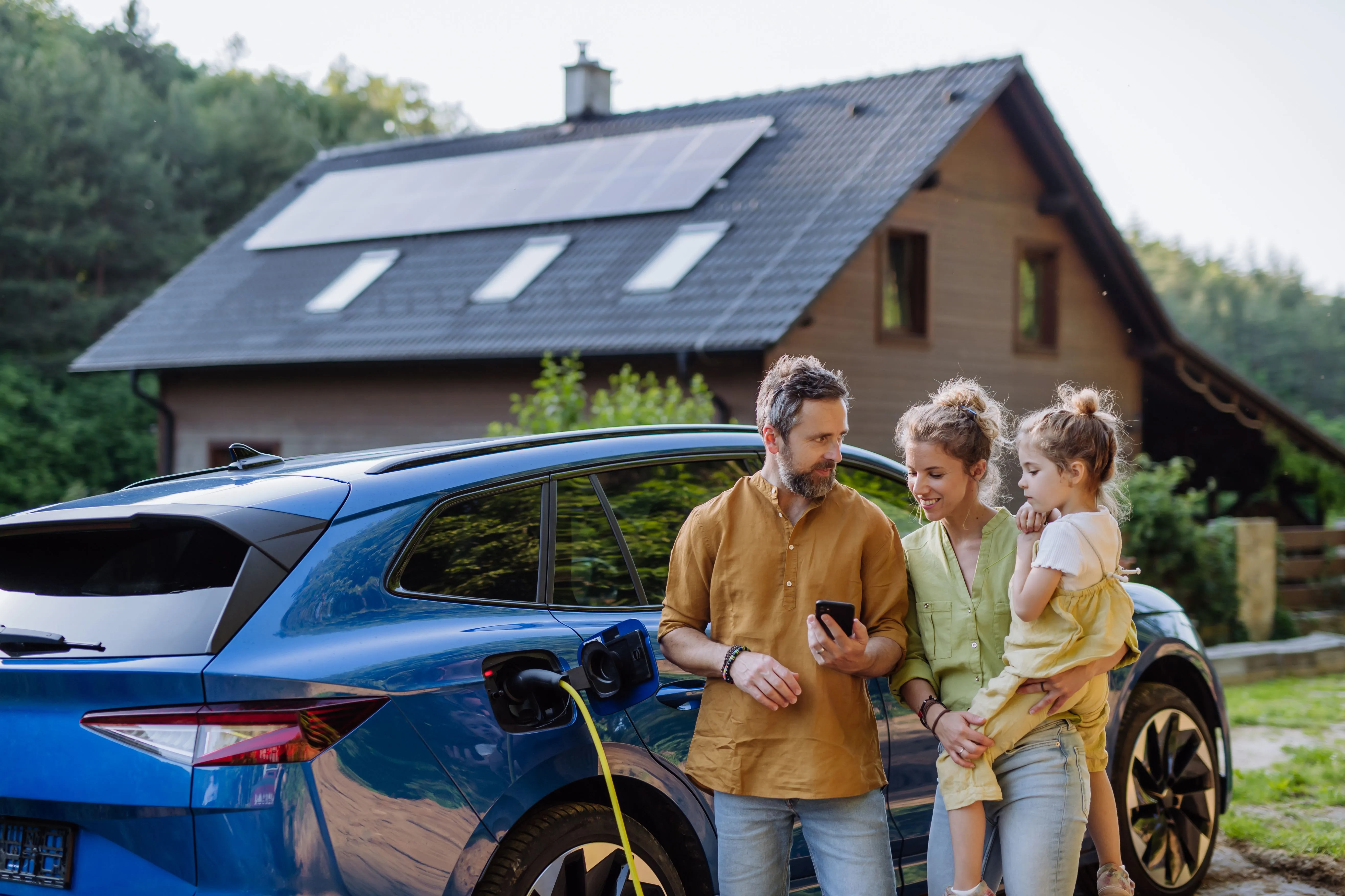 Familie mit Kind lädt vor ihrem Haus mit Photovoltaikanlage ihr Elektroauto an der heimischen Ladestation.