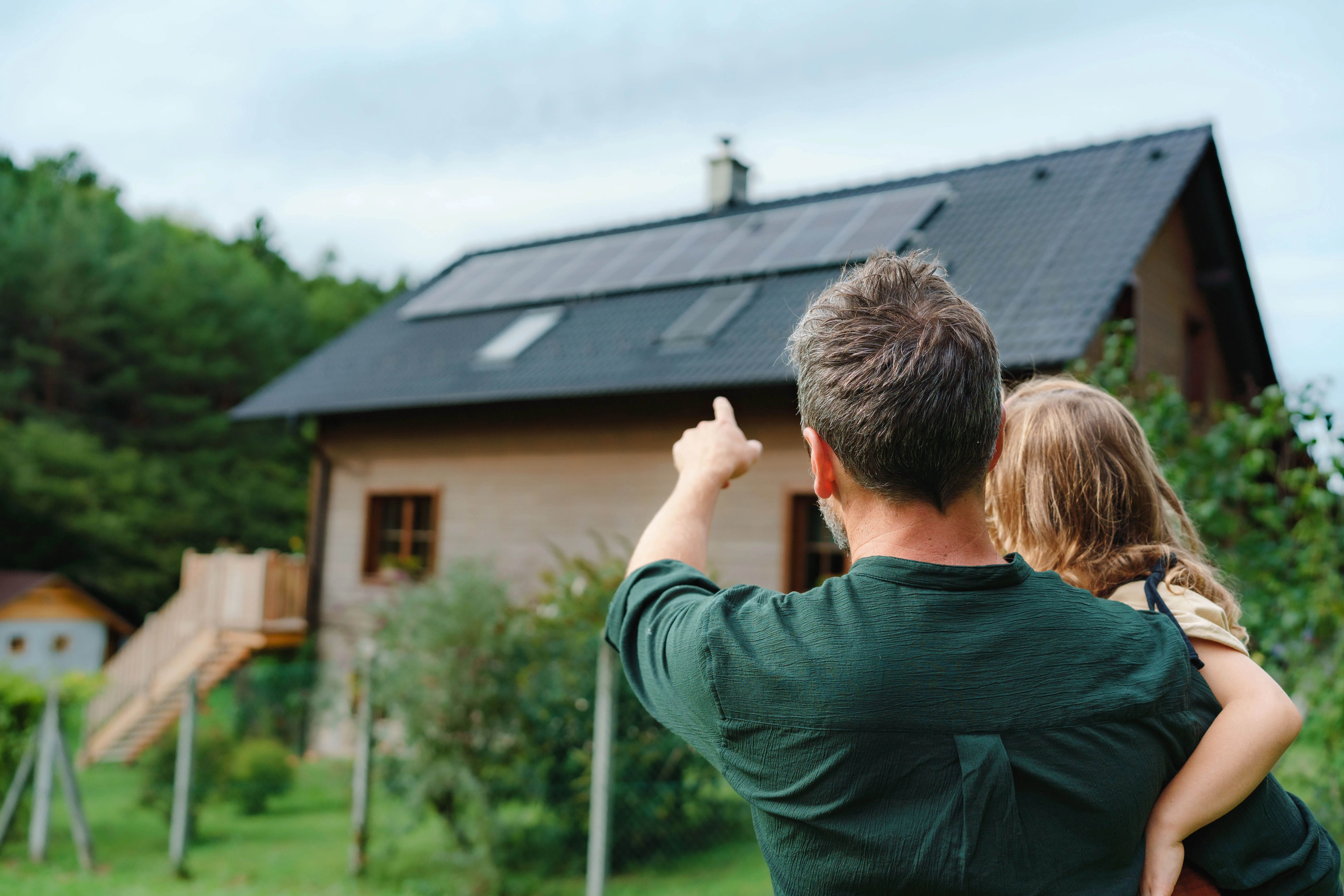 Vater mit Kind auf dem Arm zeigt auf das Photovoltaikdach ihres Hauses.