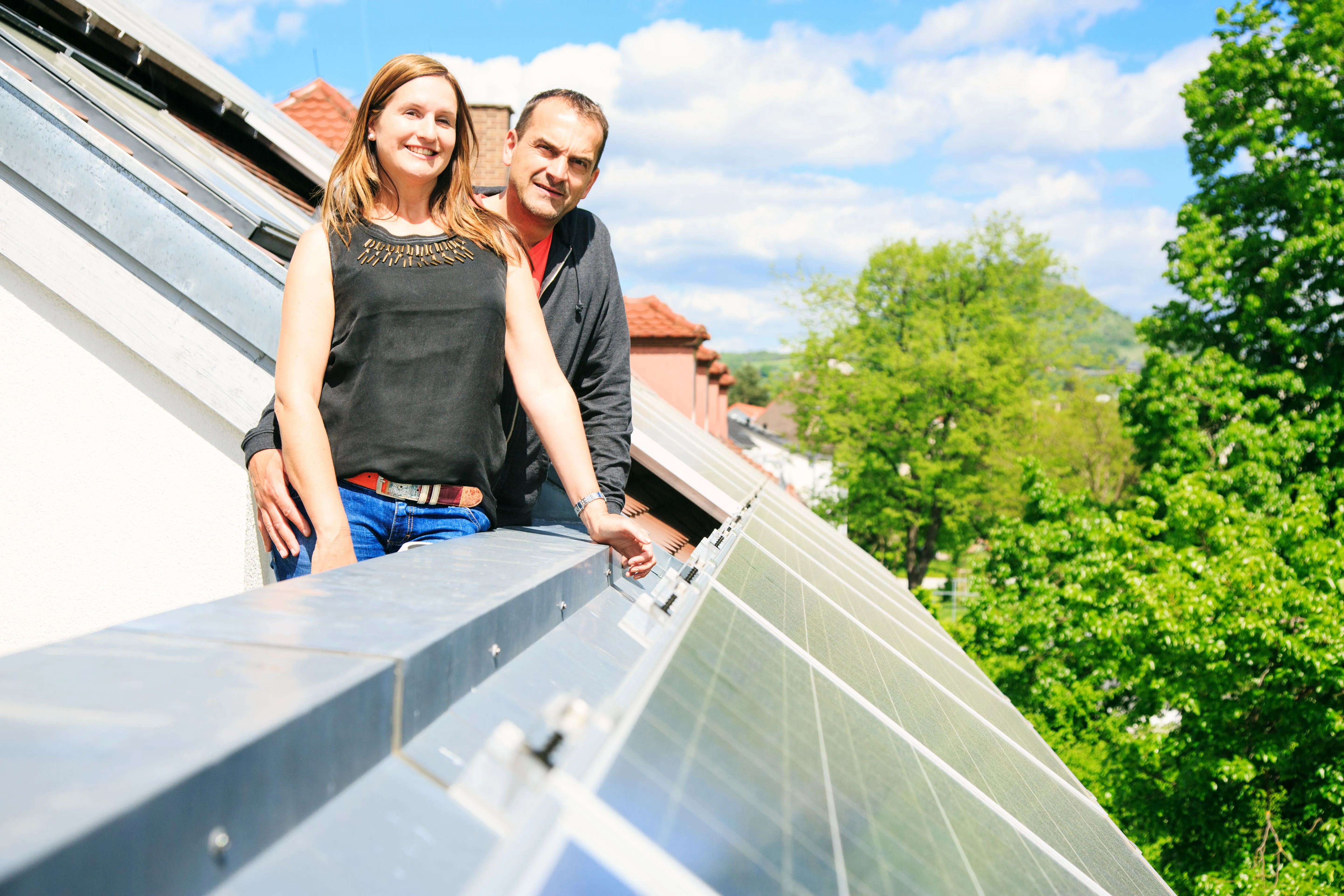 Paar steht lächelnd an einer Dachterrasse neben den Photovoltaikmodulen ihres Hauses.