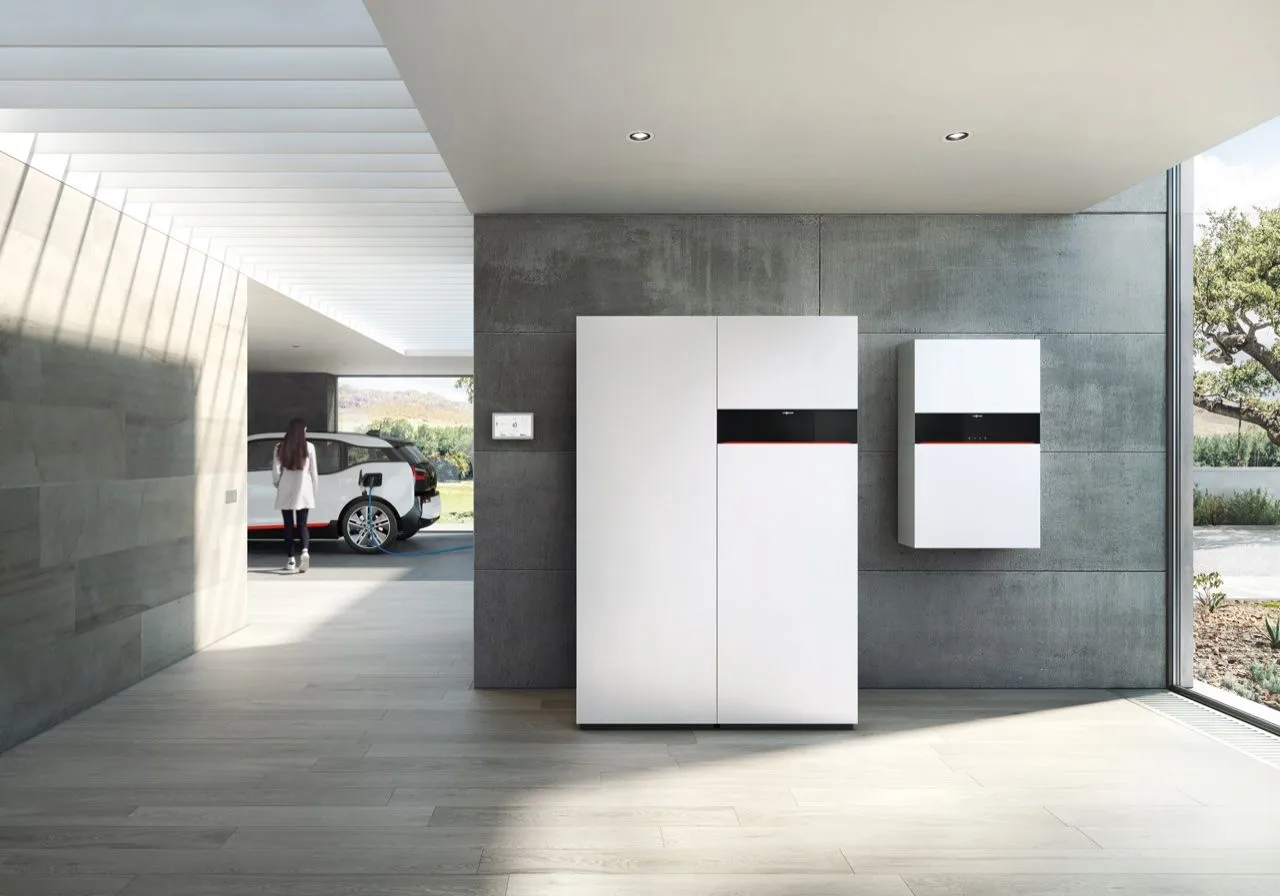 Moderner Wohnraum mit weißem Stromspeicher und Energiezentrale; im Hintergrund lädt eine Frau ihr Elektroauto.